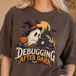 Può includere: Una t-shirt grigio scuro con la grafica di un fantasma che indossa un cappello da strega mentre usa un portatile. Il disegno include una zucca, una casa stregata, candele e il testo "DEBUGGING AFTER DARK" in un carattere audace.