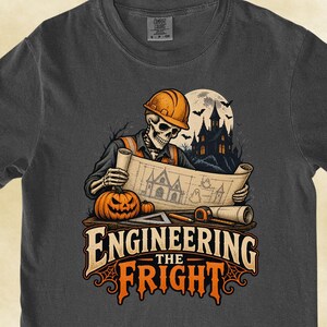 May include: A dark gray t-shirt featuring a graphic of a skeleton wearing a construction hard hat, holding a blueprint of a haunted house. A jack-o'-lantern, a pencil, and a tape measure sit nearby. The text reads Engineering the Fright.