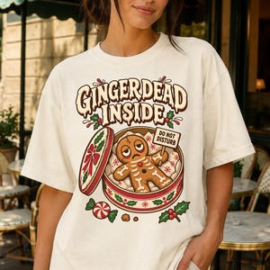 May include: An off-white t-shirt featuring a graphic of a gingerbread person inside a round tin with the text "GINGERDEAD INSIDE" and "DO NOT DISTURB" on a small sign. The design includes holly leaves, red berries, and a peppermint candy.