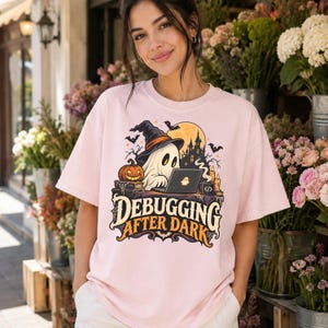 Może przedstawiać: Jasnor&oacute;żowy t-shirt z grafiką ducha w kapeluszu czarownicy, siedzącego przy biurku z laptopem, dynią i świecami. Napis "DEBUGGING AFTER DARK" wykonany jest stylizowaną czcionką, w tle widoczne są nietoperze oraz zarys zamku.