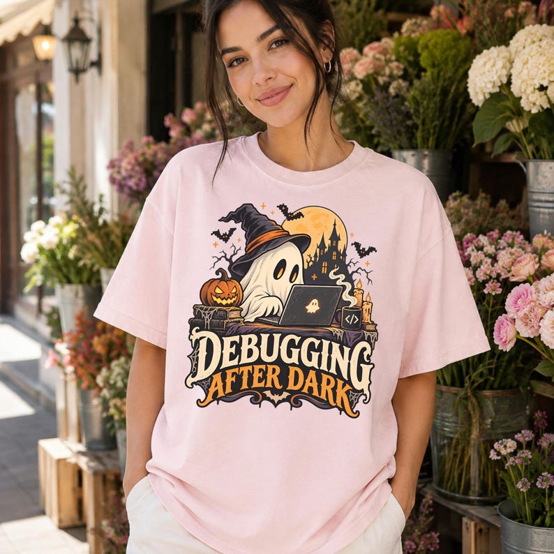 Może przedstawiać: Jasnor&oacute;żowy t-shirt z grafiką ducha w kapeluszu czarownicy, siedzącego przy biurku z laptopem, dynią i świecami. Napis "DEBUGGING AFTER DARK" wykonany jest stylizowaną czcionką, w tle widoczne są nietoperze oraz zarys zamku.