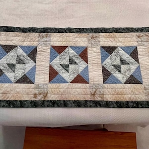 May include: A rectangular quilted table runner featuring three square designs. Each square has a central diamond pattern in blue, brown, and cream colors, framed by a light beige background and a dark green border.