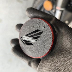 Puede incluir: Un emblema de motocicleta negro y rojo con el logotipo del ala de Honda. El emblema redondo tiene una superficie texturizada y un borde rojo. El logotipo del ala de Honda está recortado en el centro.