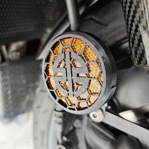 Può includere: Un indicatore di direzione per moto nero con un motivo a nido d'ape e una lente arancione. L'indicatore ha un design decorativo al centro ed è montato su una moto.