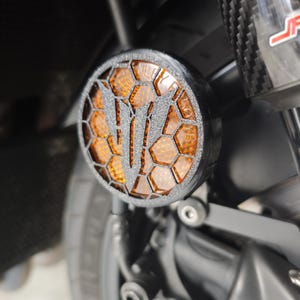 Peut inclure: Un phare de moto noir avec un motif en nid d'abeille et un "V" stylisé au centre. La lentille ambrée est intégrée dans un cadre circulaire, monté sur la suspension avant d'une moto.