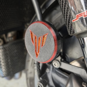 Może przedstawiać: Część motocykla z czarno-czerwonym wzorem. Okrągła część ma teksturowaną szarą powierzchnię z czerwoną obwódką i stylizowanym pomarańczowym logo. Część jest zamontowana na motocyklu.