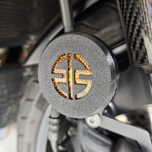 Può includere: Primo piano di un componente motociclistico. Il componente rotondo e strutturato, di colore nero, presenta un design ritagliato che si illumina con una luce arancione. La ruota della moto e altre parti sono visibili sullo sfondo.