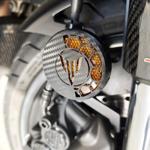 Puede incluir: Primer plano de un componente de motocicleta. Una luz redonda con textura de fibra de carbono, patrón de panal y logotipo central emite un brillo naranja. Montada en un soporte negro cerca de la rueda.