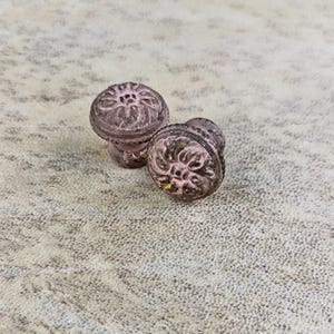 Op de afbeelding: Twee ronde metalen kastknoppen met een verweerde, vervaagde roze afwerking en een verhoogd, reliëf bloemmotief op de ronde voorzijde. Het beslag heeft een versleten textuur met zichtbare donkere metalen ondertonen en een cilindrische basis.