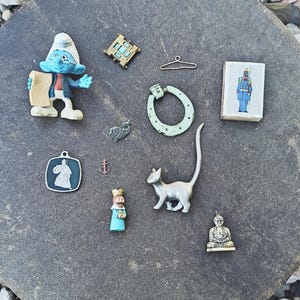 Op de afbeelding: Een verzameling kleine voorwerpen waaronder een blauw Smurf-figuurtje, een goudkleurig vierkant frame, een draadkleerhanger, een luciferdoosje met een soldaatillustratie, een lichtgroen hoefijzer, een visje, een anker, een kat, een koning en een Boeddhabeeldje.