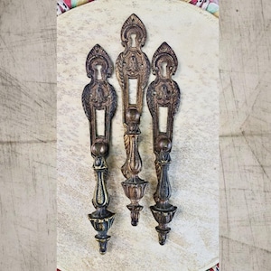 May include: Three vertical metal door plates with integrated handles, featuring a dark bronze finish and ornate, symmetrical embossed detailing. Each plate includes a keyhole at the top and a decorative, elongated handle extending downward.