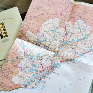 Könnte beinhalten: Eine gefaltete Papierlandkarte der Region Katalonien in Spanien mit detaillierten Straßennetzen und geografischen Bezeichnungen, präsentiert neben dem hellen Begleitheft mit dem Titel Rutas Catalanas und schwarzem sowie gelbem Text.