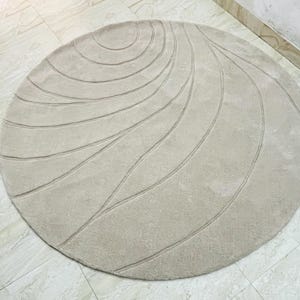 Peut inclure: Un tapis rond beige avec une surface textur&eacute;e. Le tapis pr&eacute;sente une s&eacute;rie de motifs lin&eacute;aires incurv&eacute;s dans une nuance de beige l&eacute;g&egrave;rement plus fonc&eacute;e, cr&eacute;ant un design subtil et fluide. Le tapis est pos&eacute; sur un sol carrel&eacute;.