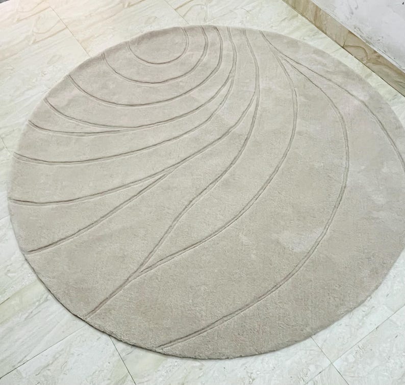 Peut inclure: Un tapis rond beige avec une surface textur&eacute;e. Le tapis pr&eacute;sente une s&eacute;rie de motifs lin&eacute;aires incurv&eacute;s dans une nuance de beige l&eacute;g&egrave;rement plus fonc&eacute;e, cr&eacute;ant un design subtil et fluide. Le tapis est pos&eacute; sur un sol carrel&eacute;.