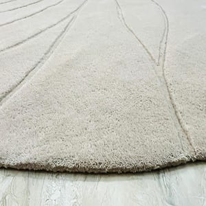 Peut inclure: Gros plan sur un tapis beige et rond avec une surface textur&eacute;e. Le tapis pr&eacute;sente un motif de lignes courbes rayonnant du centre. Le bord du tapis a une bordure plus fonc&eacute;e et effiloch&eacute;e.