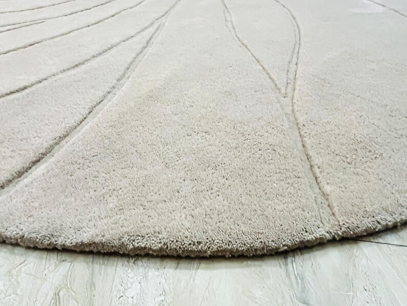 Peut inclure: Gros plan sur un tapis beige et rond avec une surface textur&eacute;e. Le tapis pr&eacute;sente un motif de lignes courbes rayonnant du centre. Le bord du tapis a une bordure plus fonc&eacute;e et effiloch&eacute;e.