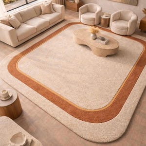 May include: A large, rectangular area rug with rounded corners. The rug features a cream-colored center bordered by a wide band of burnt orange and a thin, light-colored accent. The rug is in a living room setting with neutral-toned furniture.