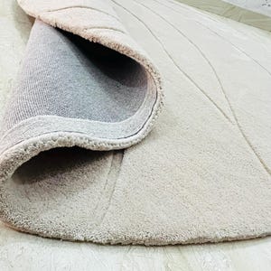 Peut inclure: Tapis rond beige avec une surface textur&eacute;e et un bord enroul&eacute;. Le tapis pr&eacute;sente un motif de lignes courbes et un dos gris. Le tapis est pos&eacute; sur un sol carrel&eacute; de couleur claire.