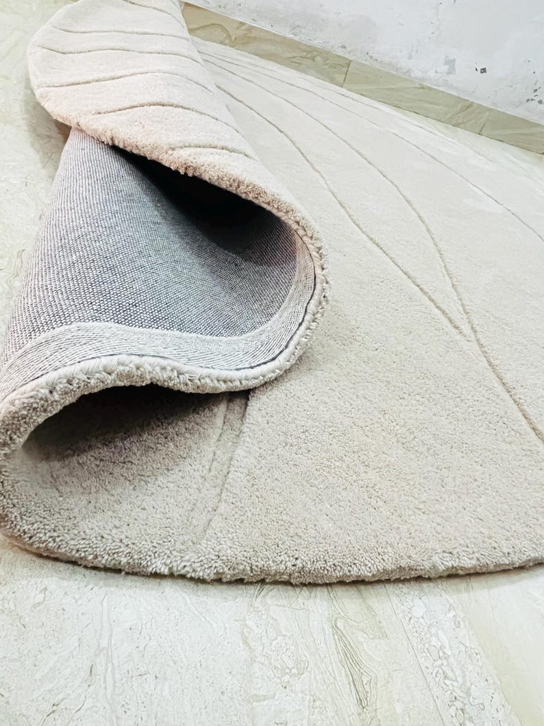 Peut inclure: Tapis rond beige avec une surface textur&eacute;e et un bord enroul&eacute;. Le tapis pr&eacute;sente un motif de lignes courbes et un dos gris. Le tapis est pos&eacute; sur un sol carrel&eacute; de couleur claire.