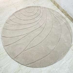 Peut inclure: Un tapis rond beige avec un motif lin&eacute;aire et tourbillonnant. Le design du tapis pr&eacute;sente des lignes courbes rayonnant du centre, cr&eacute;ant un effet textur&eacute;. Le tapis est pos&eacute; sur un sol carrel&eacute;.