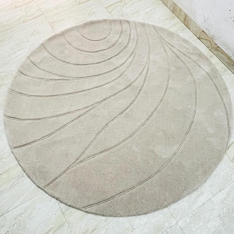Peut inclure: Un tapis rond beige avec un motif lin&eacute;aire et tourbillonnant. Le design du tapis pr&eacute;sente des lignes courbes rayonnant du centre, cr&eacute;ant un effet textur&eacute;. Le tapis est pos&eacute; sur un sol carrel&eacute;.