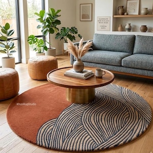 Può includere: Un tappeto rotondo con un design bicolore. Una metà è color ruggine, l'altra presenta uno sfondo beige con linee curve blu navy. Un tavolino in legno con base in ottone è appoggiato sul tappeto. La stanza comprende un divano grigio, piante e pouf in pelle.