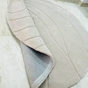 Peut inclure: Tapis beige enroul&eacute; avec un motif incurv&eacute; et textur&eacute;. La surface du tapis pr&eacute;sente des lignes parall&egrave;les en relief, cr&eacute;ant un motif en forme de vagues. Le dessous du tapis est visible, montrant un envers gris.