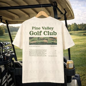 May include: Cream-colored t-shirt with "Pine Valley Golf Club" printed in green. The shirt features a landscape image and text about the 7th hole. A golf cart and golf clubs are in the background.