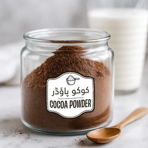 Może przedstawiać: Przezroczysty szklany słoik wypełniony ciemnobrązowym kakao w proszku. Biała etykieta na słoiku zawiera napis "COCOA POWDER" w języku angielskim i innym języku. W pobliżu znajduje się drewniana łyżka i szklanka mleka.