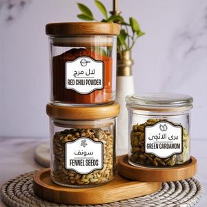 May include: Three glass spice jars with wooden lids and labels. The jars contain red chili powder, fennel seeds, and green cardamom. The jars are arranged on a wooden tray, with a white vase and greenery in the background.