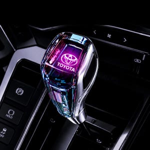 May include: A gear shift knob featuring a multi-faceted, translucent crystal-like design with iridescent blue and purple hues. The top surface displays the white Toyota logo and text. The base is metallic silver, mounted on a black car console.