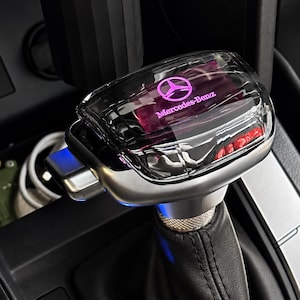 May include: A gear shift knob featuring a clear, transparent top housing with an illuminated pink Mercedes-Benz logo and text. The base is constructed from silver-toned metal and black leather, with a textured metallic collar below the clear top.