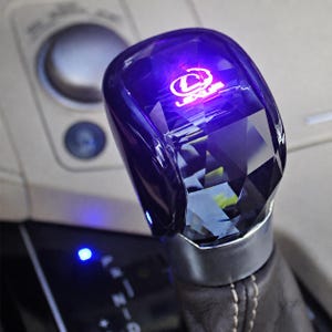 Op de afbeelding: Een versnellingspookknop in kristalstijl met een gefacetteerd oppervlak en een gloeiend paars LED-licht. De bovenkant van de knop is voorzien van het verlichte witte Lexus-logo. De basis is van zilverkleurig metaal en verbonden met een donkere lederen hoes.