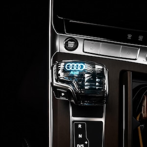 以下が含まれることがあります： Audi車両用の透明なシフトノブ。青く光るAudiのロゴとテキストが特徴です。シフトノブはダークな多面体デザインで、側面にPボタンがあり、現代的な車のセンターコンソールに配置されています。
