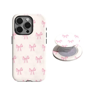 Op de afbeelding: Een witte telefoonhoes en bijpassende telefoonhouder met een herhalend patroon van roze aquarel strikken. De telefoonhoes heeft een zwarte cameralens en zwarte zijknoppen. De telefoonhouder is wit met een roze strikpatroon.