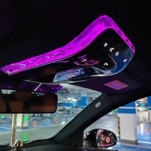 May include: An overhead car console featuring a glossy black finish with integrated purple ambient lighting. The console includes several illuminated control buttons, a small digital display, and a rearview mirror positioned below it.