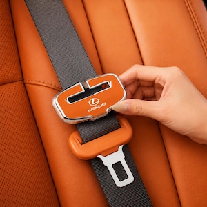 May include: A dark gray seat belt featuring a custom orange and chrome buckle cover with the Lexus logo in white text. The buckle cover is rectangular with rounded edges and is positioned over the standard metal seat belt release mechanism.
