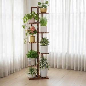 May include: A tall, dark brown wooden plant stand with multiple shelves, displaying various potted plants and flowers. The plants are in different sized pots and containers, with a variety of green foliage and colorful blooms. The stand is positioned in front of a sheer white curtain.