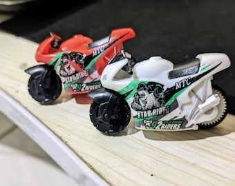 May include: Two toy motorcycles, one red and one white, are positioned side-by-side. Each motorcycle features black wheels, a streamlined design, and graphics that include the text "STAR-RIDER" and "92 RIDERS". The motorcycles are on a light-colored surface.