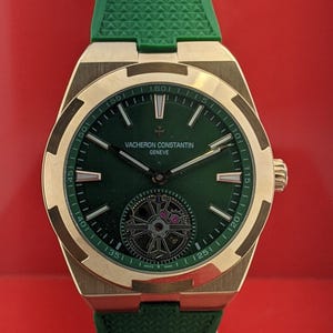 May include: A gold-cased wristwatch with a green dial and strap. The watch face displays the brand name "Vacheron Constantin Geneve". The watch is presented on a clear stand against a red backdrop.