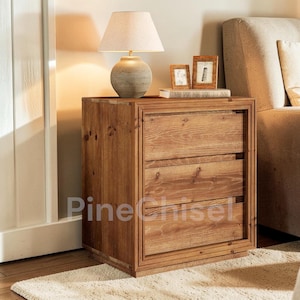May include: A wooden nightstand with three drawers, a lamp, and two small picture frames. The nightstand is made of light brown wood and has a rectangular shape. The lamp has a beige lampshade and a round, textured base. The picture frames are brown.