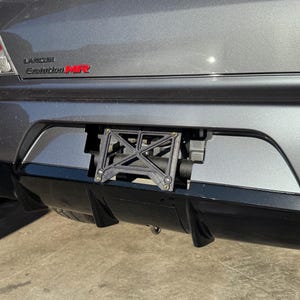 May include: A close-up of the rear of a gray car, featuring a black license plate holder. The car has a black bumper and the text "Lancer Evolution MR" in red. The license plate holder is a black, geometric design.