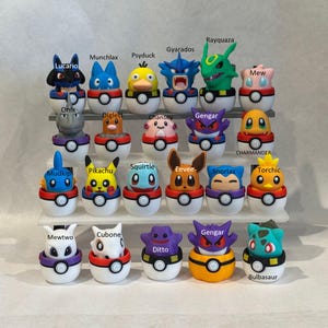 Puede incluir: Una colección de veinte pequeñas figuras de personajes coloridos, cada una emergiendo de una base esférica blanca y de color. Las figuras incluyen a Lucario, Munchlax, Psyduck, Gyarados, Rayquaza, Mew, Onix, Diglett, Chansey, Gengar, Charmander, Mudkip, Pikachu, Squirtle, Eevee, Snorlax, Torchic, Mewtwo, Cubone, Ditto y Bulbasaur, dispuestas en un soporte de exhibición escalonado.