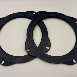 May include: Two black speaker mounting rings with multiple screw holes. The rings are circular with a slightly textured surface and are designed to fit around a speaker. The rings are made of a durable material.
