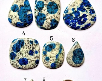 May include: A collection of eight polished k2 azurite gemstone cabochons in various shapes, including teardrop, oval, and rectangular. The stones feature a white base with blue azurite inclusions and black veining. A US dollar coin is included for size reference.