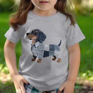 May include: A heather gray short-sleeve t-shirt featuring a graphic of a dachshund dog wearing a patchwork denim jacket. The dog has black and tan fur, and the jacket is composed of various shades of blue denim fabric with visible stitching.