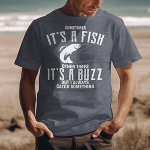 Könnte beinhalten: Ein dunkelgraues T-Shirt mit weißem Text und einer Fischgrafik. Der Text lautet: "Sometimes It's A Fish, Other Times It's A Buzz, But I Always Catch Something". Das Shirt hat kurze Ärmel.