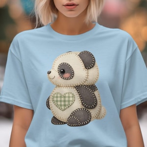 May include: Light blue t-shirt featuring a cute panda bear design. The panda has a cream-colored body, black accents, and a green plaid heart. The design is stitched with a cross-stitch pattern.