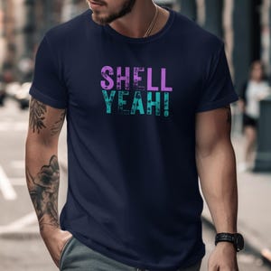 Puede incluir: Camiseta azul marino con el texto "SHELL YEAH!" en una fuente desgastada y multicolor. El texto es en tonos morados y turquesas. La camiseta es de cuello redondo.