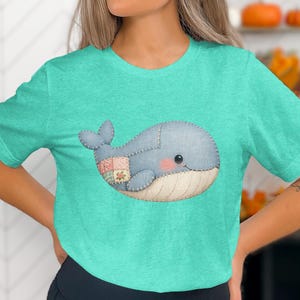 Puede incluir: Una camiseta turquesa con un diseño de ballena caprichoso. La ballena es azul claro con un detalle de patchwork y una cara sonriente. El diseño está centrado en la camiseta, creando una estética encantadora. La camiseta es de material suave.